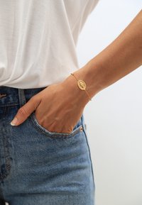 Guldring med en oval berlock, med en texturerad yta. Bärs på en hand nära en ficka på blå jeans.
