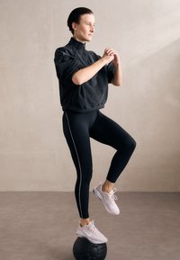 Giacca nera in pile con mezza zip e polsini elastici, abbinata a leggings neri con strisce laterali bianche. Scarpe sportive rosa chiaro.