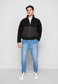 Musta fleece-takki, jossa on ristiriitainen musta nylon-paneeli, vetoketjukiinnitys ja kaksi etutaskua, yhdistettynä vaaleansinisiin farkkuihin ja valkoisiin tennareihin.