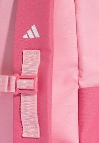 Ružový polyesterový batoh s textúrovanou tkaninou, nastaviteľným popruhom a bielym logom značky na boku. Má zabezpečené zapínanie na pracku.