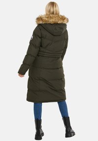 Lunga giacca trapuntata verde oliva con cappuccio foderato in pelliccia, dotata di vita cinturata e tasche laterali. Indossata sopra jeans blu e stivali neri.