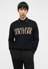 Άνδρας που φοράει μαύρο πουλόβερ Versace Jeans Couture πάνω από μαύρο πουκάμισο με γιακά, με τα χέρια στις τσέπες μαύρου παντελονιού, σε απλό φόντο.