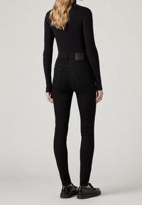 Femme portant un haut noir côtelé à manches longues, un jean skinny noir taille haute et des mocassins noirs, debout devant un fond uni.