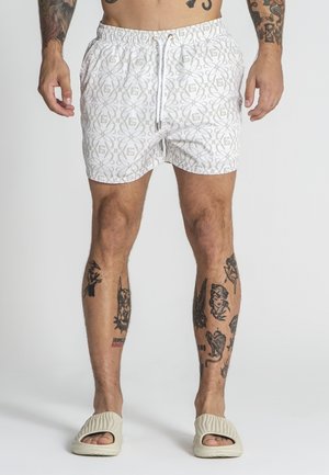 Weiße Badeshorts mit einem geometrischen grauen Muster. Verfügt über einen elastischen Bund mit Kordelzug. Kombiniert mit strukturierten beigen Sandalen.