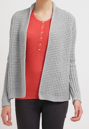 Kvinde iført en lysegrå åben strikket cardigan over en rød waffle-strikket top med knapper og mørke bukser, stående foran en ensfarvet baggrund.