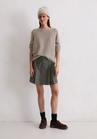 Beige gebreide trui, grijs geruite geplooide rok, bruine loafers met contrasterende sokken, gedragen tegen een textuurvolle witte achtergrond.