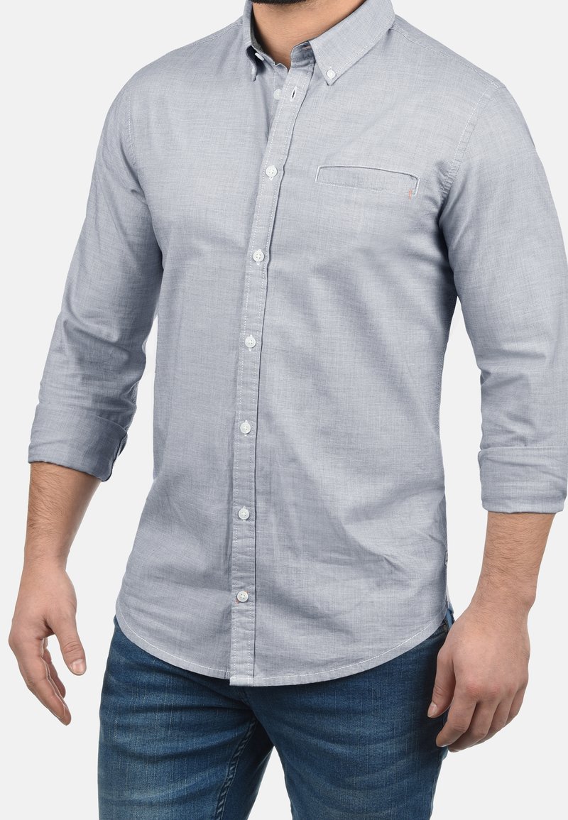 Camicia azzurro chiaro con bottoni in tessuto strutturato, dotata di una piccola tasca sul petto e maniche arrotolate. Abbinata a jeans blu scuro.