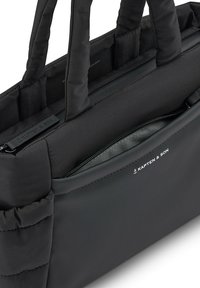 Bolsa de tote de tela negra con asas acolchadas, que cuenta con un bolsillo exterior con cremallera y un logo sutil. Textura suave con un diseño estructurado.