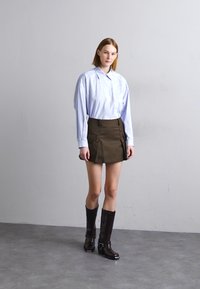 Ανοιχτό μπλε πουκάμισο μακρύ μανίκι με κοφτή κολάρο, συνδυασμένο με καφέ μίνι φούστα με τσέπες, και καφέ μπότες ως το γόνατο με μεταλλικές λεπτομέρειες.