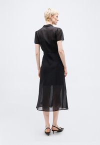 Άτομο με κοντά ξανθά μαλλιά που φοράει ένα διάφανο μαύρο μέτριο φόρεμα με κοντά μανίκια και μαύρα παπούτσια με λουράκι στη φτέρνα, στέκεται μπροστά σε μονόχρωμο φόντο.