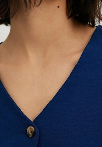 Nahaufnahme eines Halses und des oberen Brustbereichs einer Person, die ein blaues V-Ausschnittoberteil mit einem runden braunen Knopf trägt. Kurze dunkle Haare sind sichtbar.