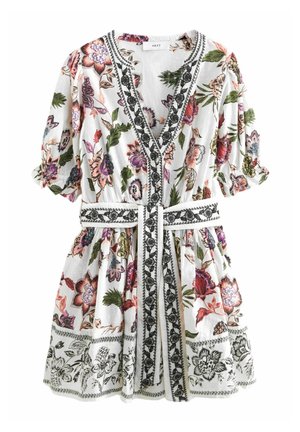 Mujer con vestido blanco floreado con flores rosas y moradas, ribete bordado en blanco y negro, escote en V y cinturón anudado en la cintura.
