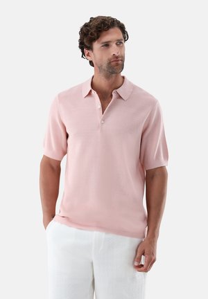 Mann mit lockigem Haar, der ein hellrosa kurzärmeliges Polohemd und weiße Hose trägt, vor einem einfarbigen Hintergrund stehend.