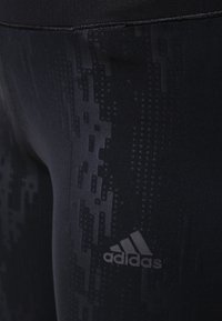 Las mallas deportivas negras presentan un patrón texturizado y un logo de Adidas gris en la parte inferior de la pierna. El material parece suave y elástico.