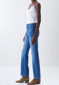 Jeans de mezclilla azul con piernas acampanadas, cintura alta y bolsillos delanteros. Se combinan con una camiseta de tirantes blanca de canalé y botas marrones hasta el tobillo.