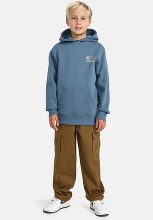 Blauwe hoodie met een voorzak en logo-detail, gecombineerd met bruine cargobroeken en witte sneakers met groene accenten.