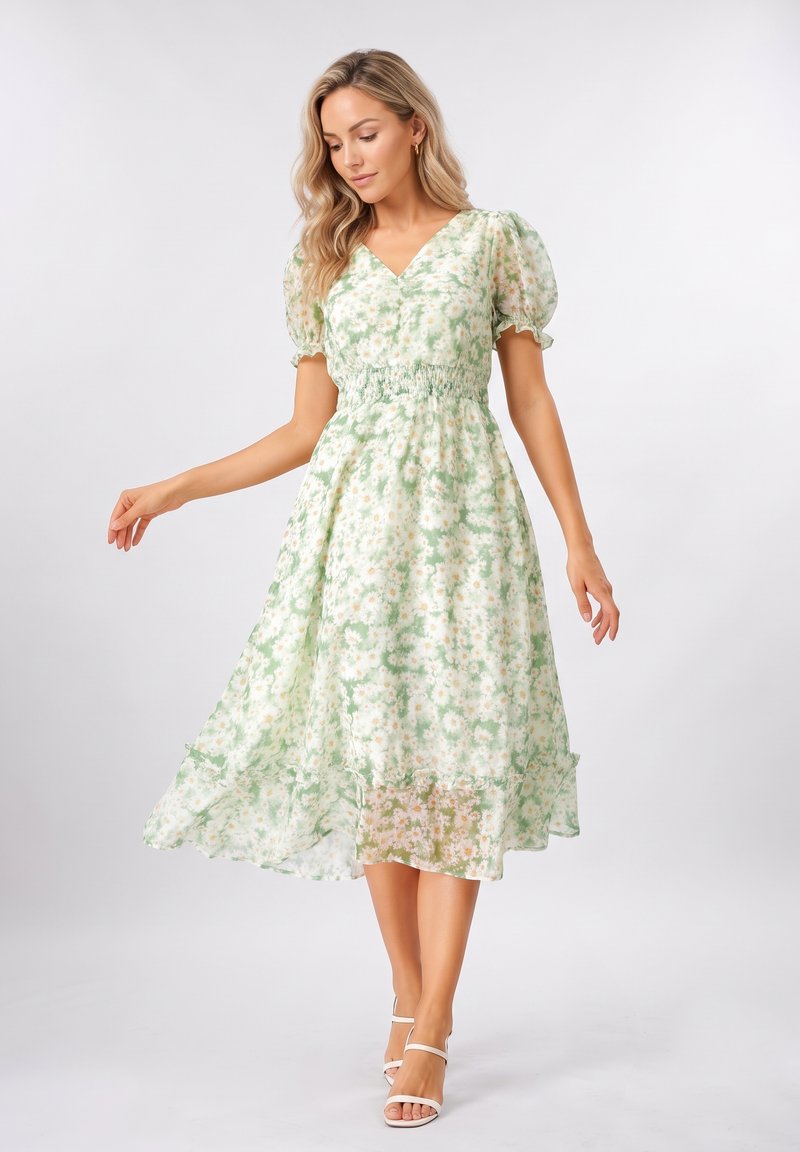 Femme portant une robe midi fleurie vert clair avec manches bouffantes et des talons blancs ouverts, debout devant un fond gris uni.
