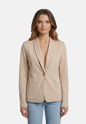 Femme aux cheveux mi-longs portant un blazer beige à un bouton et un jean bleu clair, debout devant un fond clair uni.