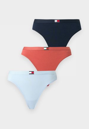 Mladá žena v bielom bralette Tommy Hilfiger a tmavomodrom spodnom prádle stojí na jednoliatom svetlom pozadí s jednou rukou na bokoch.