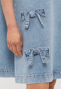 Vestido de ganga azul claro com dois pequenos laços em tecido coordenado, colocados na área inferior da frente, com detalhes de costura visíveis.