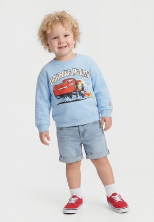 Copil mic zâmbitor cu păr creț blond, purtând un hanorac albastru deschis cu Lightning McQueen, pantaloni scurți din denim, șosete albe și adidași roșii.