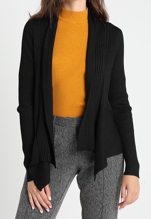 Mujer con una blusa de cuello alto de canalé color mostaza, cárdigan negro abierto al frente y pantalones grises texturizados, de pie con las manos relajadas.