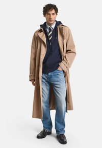 Beige trench coat over a navy hooded sweatshirt, paired with light blue jeans and black leather shoes. Casual layered outfit.