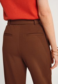 Bruna skräddarsydda byxor med två bakfickor, i en texturerad tyg och med hög midja. Outfitten inkluderar en röd topp och guldaccessoarer.