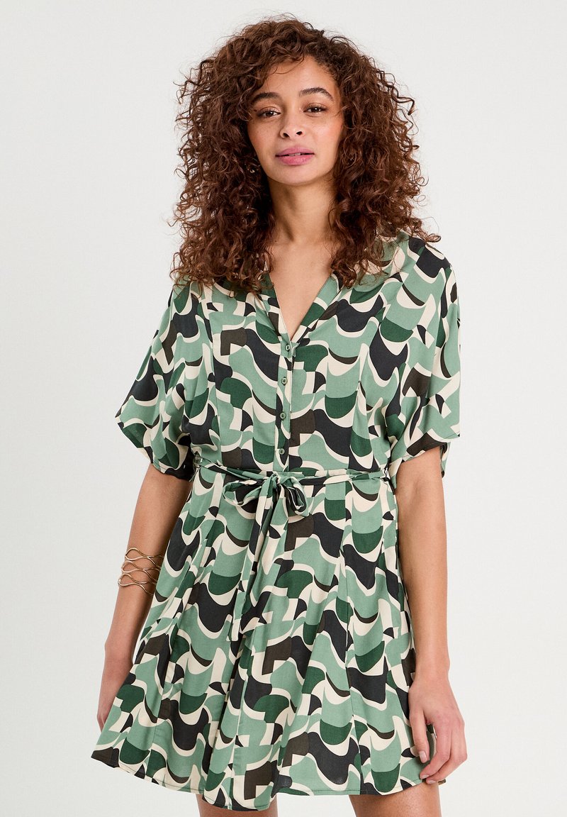 Robe à manches courtes avec un bouton en vert, noir et crème avec des motifs en vagues, cintrée à la taille avec une ceinture en tissu assortie. Texture douce.