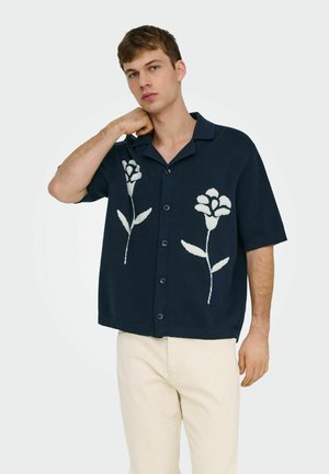 Młody mężczyzna w granatowej koszuli z krótkim rękawem zapinanej na guziki z białymi kwiatowymi wzorami oraz jasnobeżowych spodniach, pozujący na jednolitym tle.