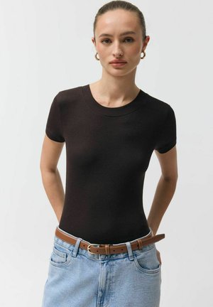 Kobieta z włosami związanymi do tyłu, nosząca złote kolczyki-koła, czarną bluzkę z krótkim rękawem, brązowy pasek i jasnoniebieskie jeansy na jednolitym tle.