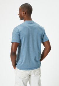 Blaues Baumwoll-T-Shirt mit Rundhalsausschnitt und kurzen Ärmeln, das eine glatte Textur und eine lockere Passform aufweist, von hinten gezeigt.