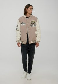 Męska beżowa i kremowa kurtka varsity z haftowanym wzorem tygrysa, zapięcie na napy, paski na mankietach, w zestawie z czarnymi dżinsami i białymi sneakerami.