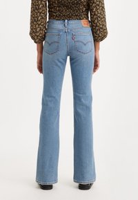 Femme portant un jean flare taille haute bleu clair et un chemisier à manches longues sombre à motifs floraux, debout, de dos, devant un fond uni.