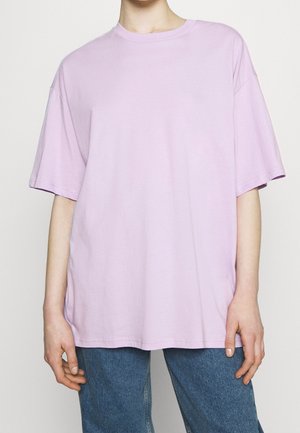 Osoba ubrana w luźną, pastelowo-fioletową koszulkę z krótkim rękawem i niebieskie dżinsy, stojąca z ramionami swobodnie opadającymi wzdłuż ciała.