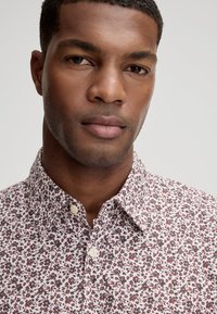 Hemd met knopen, voorzien van een witte achtergrond met kleine rode en zwarte bloemenpatronen, een gestructureerde kraag en zichtbare knoopdetails.