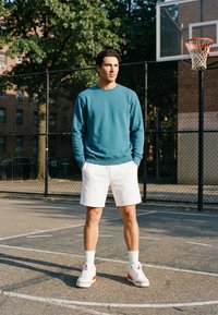 Jeune homme debout sur un terrain de basket en plein air portant un sweat-shirt sarcelle, un short blanc et des baskets près du panier de basket et de la clôture.