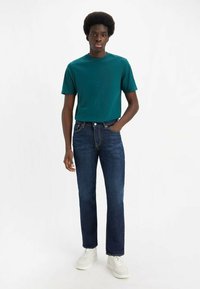 T-shirt di cotone color teal abbinata a jeans in denim blu scuro, con taglio dritto e classico design a cinque tasche, indossata con sneakers bianche.