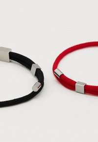 Kaksi rannekorua, yksi musta ja yksi punainen, valmistettu punotusta nyöristä, joissa metalliset neliönmuotoiset koristeet ja sulkimet. Yksinkertainen, moderni muotoilu, jossa kontrastivärit.