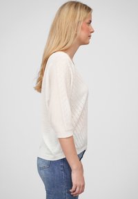 Hvit strikkegenser med løs passform, med kort erme, teksturert diamantmønster og avrundet kant. Kombinert med blå jeans.
