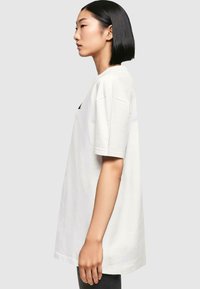 White cotton T-shirt with short sleeves, loose fit, ribbed crew neck, and a subtle logo on the side. Smooth texture.