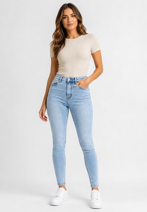 Vrouw staand, draag een beige top met korte mouwen, lichtblauwe high-waisted skinny jeans en witte sneakers tegen een witte achtergrond.