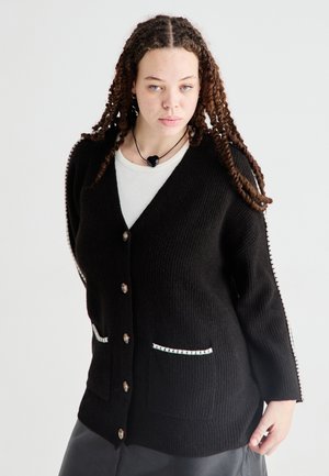 Schwarze gerippte Strickjacke mit V-Ausschnitt, Knopfverschluss und zwei vorderen Taschen. Mit weißen Nähten an den Seiten und dekorativen Knöpfen.