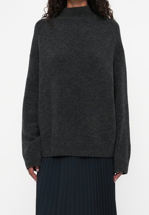 Kobieta ubrana w ciemnoszary, długi sweter dzianinowy z półgolfem i czarną plisowaną spódnicę, stojąca na tle jednolitego białego tła.