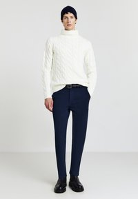 Cremiger, kabelstrick-gestrickter Rollkragenpullover mit gerippten Bündchen, kombiniert mit dunkelblauen, maßgeschneiderten Hosen. Das Model trägt schwarze Stiefeletten.