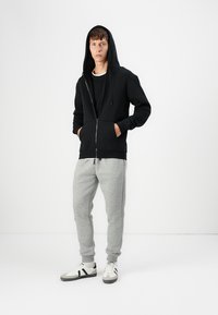Sudadera con cremallera negra, con bolsillos frontales y capucha con cordón, combinada con pantalones de chándal grises y zapatillas blancas con acentos negros.