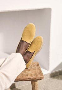 Yellow suede espadrilles with a slip-on design, accented by a braided jute sole. Placed on a wooden stool with visible texture.