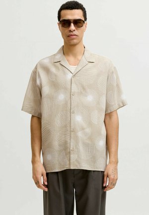 Mann trägt braune Sonnenbrille und beige kurzärmliges Hemd mit weißen kreisförmigen Mustern und dunkelbrauner Hose.