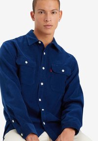 Camisa de pana azul marino con un diseño de botones, que presenta dos bolsillos en el pecho, botones blancos y una superficie ligeramente texturada.