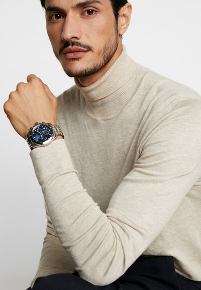 Pull en cachemire gris pour homme avec col roulé côtelé, porté avec une montre chronographe en acier inoxydable argenté dotée d'un cadran bleu et de trois sous-cadrans.